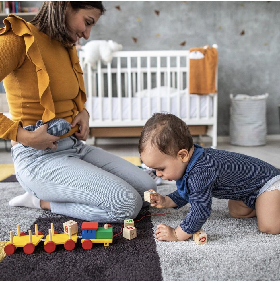 Woman and baby playing with a wooden train on a rug in a nursery room.